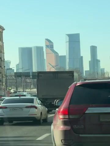 The video depicts a traffic jam on a sunny day, with a view of the Moscow International Business Center skyline in the background. Several cars, including a white sedan and a maroon Jeep Grand Cherokee, are visible in the foreground. The cars are stationary or moving very slowly. The sky is clear and blue. The buildings of the business center are tall and modern, with various shapes and glass exteriors. A large truck is also present among the cars.
