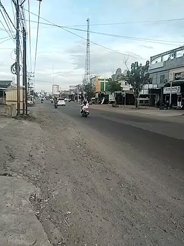 The video shows a shot of a nearly empty street in what appears to be a small town in Southeast Asia. A few motorbikes are on the road, and several shops and stalls line the side of the street. The sky is overcast and a cell tower is visible in the distance. There is a large tree beside a small building. A few parked motorcycles are observed near storefronts. The video appears to be taken from the side of the road, looking toward the buildings.