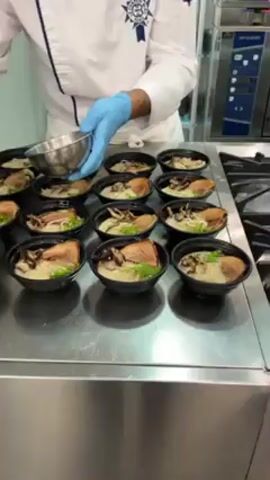 In a bright, well-lit professional kitchen, a chef wearing a white coat and blue gloves meticulously prepares multiple servings of ramen. The chef is seen pouring ingredients from a stainless steel bowl into each of the black bowls, each containing ramen noodles, broth, and what appear to be slices of meat and mushrooms.  The stainless steel counter is spotless.  A large stainless steel oven and other kitchen equipment are visible in the background, suggesting a commercial kitchen setting. The chef is focused on evenly distributing the final topping to each bowl, indicating attention to detail and precision in the preparation process.