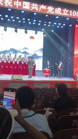 A vertical medium shot captures a choir on stage. There are over twenty singers in maroon robes with a handful of others in white shirts and black ties. A male conductor in a black suit leads them from the center of the stage. To the right of the stage are three male musicians. One plays a large red drum, another plays a flute, and another is barely visible at the very right edge of the frame. Behind the choir is a large screen with a landscape image of mountains and a red sun. Several white stage lights illuminate the stage from the top. The foreground shows several rows of empty seats with two audience members partially visible in the bottom right corner. A hand holding a cell phone recording the performance is visible at the bottom left.