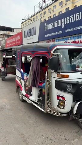 The video shows a street scene in Cambodia. Two tuk-tuks are parked at the side of the road. The tuk-tuk closest to the camera is white with red and blue accents. The words "Thailand attack first" are visible on its side. A man is sitting in the other tuk-tuk, which is red. In the background, there are buildings with signs in both English and Khmer. One sign advertises "Cambodia Premium Water." There are other vehicles parked along the street, and a few people are walking around. The sky is overcast.