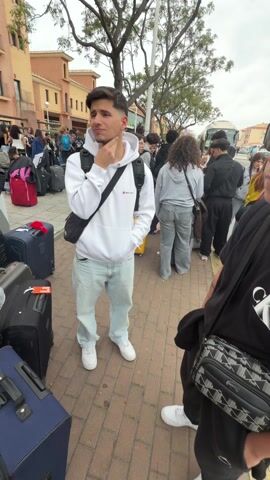 The video shows a group of people standing on a sidewalk with their luggage, waiting to board a bus. The group includes two young men in the foreground, one wearing a white hoodie and light-wash jeans, and the other wearing a black T-shirt and sunglasses. The first man has his hand on his chin, as if in thought. They are both wearing crossbody bags. The other people are lined up behind them with their suitcases. Several of them are boarding a white bus that is partially visible in the background. The environment consists of a brick sidewalk, buildings in the background, and a tree with bare branches. The scene appears to be a gathering of people preparing for transportation.