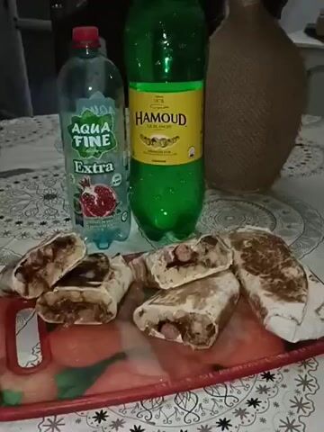 A close-up shot presents a red plastic cutting board on a table, displaying five cut shawarmas, likely containing sausage, potatoes, and sauce. Behind the board are two bottles: one of Aqua Fine Extra pomegranate-flavored water and one of Hamoud La Blanche soda. A brown, vase-like object is visible to the right. The table is covered with a white tablecloth featuring a repeating floral pattern. The shot is static with subtle camera movements.