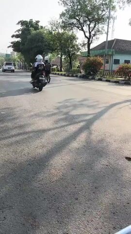 The video shows a street scene in Kludan, East Java, during daylight hours. The sun casts long shadows across the asphalt road, suggesting it is either early morning or late afternoon. The weather appears clear and sunny.

The primary subjects are two individuals riding on motorcycles. The foreground motorcycle has a rider wearing a white helmet and a dark jacket with white accents. A passenger is also visible on the back of this motorcycle. Behind them, another motorcycle and rider are partially visible. The road is paved and appears to be a public street, with a sidewalk and curb on the right side.

The setting includes a variety of trees and some buildings in the background. One building is light-colored with a green roof and appears to be a commercial or institutional building. There are also some flowering bushes along the roadside. In the distance, a white car is seen driving on the road.

The activity taking place is simply traveling on motorcycles along a street. There are no overt signs of any particular event or strong emotions. The scene is calm and ordinary. No visible text is present in the video.