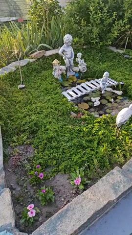The video shows a garden scene with various statues and plants. In the center, a small pond is surrounded by lush green ground cover. A white wooden bridge spans a section of the pond. Several small white duck statues are in the pond, and a larger white bird statue stands near the edge. On a raised mound behind the pond, there are three statues of cherubic figures, one appearing to be urinating. To the left of the mound, there's a statue of a figure wearing a straw hat. Small pink flowers are blooming in the foreground, planted in soil between stone borders. The lighting suggests it is either early morning or late afternoon, casting soft shadows. The overall impression is a whimsical, decorative garden setting.