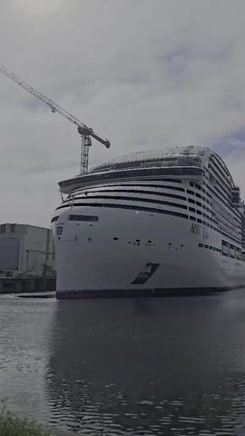 A slow-motion, low-angle exterior shot shows the bow of a massive white cruise ship docked at a harbor. The ship has multiple decks and a distinctive arched, glass-covered top deck. The MSC logo is visible on the side of the ship. A large construction crane stands behind the ship, its boom extending over the top deck. The water in the foreground is choppy, reflecting the overcast sky. A building is visible to the left of the ship.