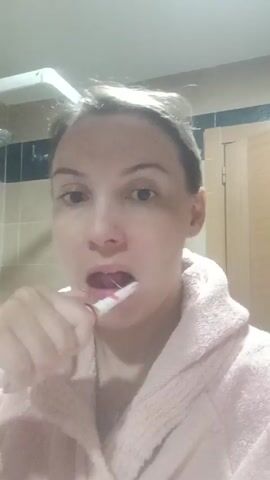 A woman in a pink bathrobe is brushing her teeth in a bathroom. She is facing the camera, and the bathroom has tiled walls, a shower, and a wooden door. She moves the toothbrush in a circular motion over her teeth. Her hair is pulled back and slightly messy.