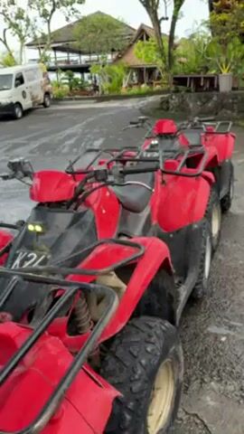 Two red ATVs are parked side by side on an asphalt surface. The ATVs are facing the same direction, with the first one slightly angled towards the camera. The front ATV has a visible license plate that appears to read "K24". Behind the ATVs, there is a white van parked on the left. In the background, there's a building with a thatched roof. Lush greenery and other buildings are also visible in the distance. The weather seems overcast.