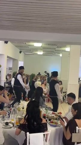 The video shows a folk dance performance in a banquet hall. Four people in traditional costumes are dancing among tables of seated guests. The men wear black caps and vests over white shirts, while the women have colored headscarves and patterned skirts. Guests are seated at tables covered with white linens and centerpieces of white flowers. Some are using their phones, while others watch the dancers. The room has a white ceiling with recessed lighting and white walls. A DJ booth with equipment is visible in the background.