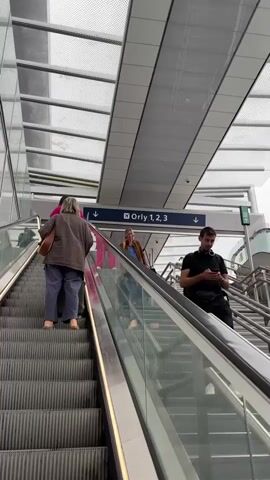 An upward-moving escalator is shown in a modern transit station. Several people are on the escalator, some moving up, others moving down. One person is on the far left of the escalator, looking ahead. Another person in pink pants is further up, also looking ahead. A woman in a brown jacket and blue top is on the descending escalator, looking towards her phone. A man in a black shirt is on the right side of the descending escalator, also looking at his phone. Above the escalators, a sign indicates "Orly 1, 2, 3" with arrows pointing to the right. The ceiling of the station features a modern design with glass and metal panels.