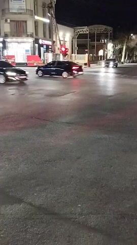 At night, several cars are driving on a wet asphalt road. The traffic light is red.  Buildings with lighted windows and signs are visible on the side of the road.  The cars include a black sedan, a white sedan, a silver sedan, and a white SUV.