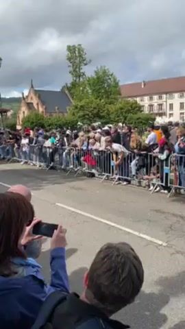 The video shows a crowd of people standing behind metal barricades, watching a blue vintage race car drive past. Many people in the crowd are holding up phones to record the event. Buildings and trees can be seen in the background, and the sky is partly cloudy.