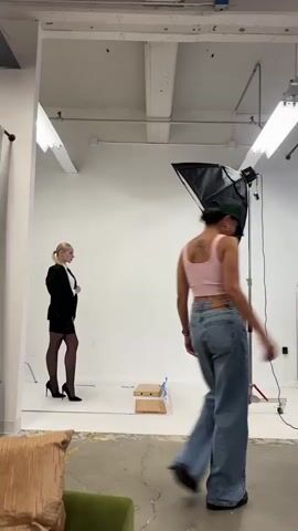The video shows a photography studio with a white backdrop. A blonde woman in a black blazer, white shirt, black skirt, black stockings, and black high heels stands on the left side of the frame. A photographer in a pink crop top and blue jeans is on the right, holding a camera and taking a picture of the blonde woman. The photographer appears to be directing the model, as they are looking at the model and holding the camera up. The studio is equipped with professional lighting equipment, including a large softbox on a stand, and overhead fluorescent lights. There is also a large fan visible in the foreground on the right side of the frame. The floor is a concrete surface, and part of a green couch is visible in the bottom left corner. The overall atmosphere suggests a professional photo shoot.