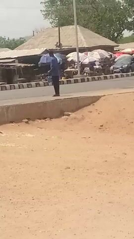 The video shows a roadside scene in what appears to be a developing country. There's a street lined with vendors selling goods under umbrellas, a man standing on the side of the road, a parked vehicle, and trees in the background. There are also streetlights. The foreground consists of dirt ground. The scene seems ordinary, depicting everyday life.
