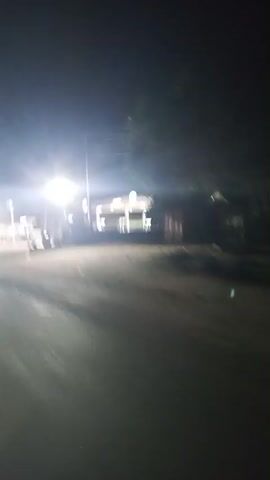 The video shows a gas station at night. The station is brightly lit with white lights, and there are gas pumps under a canopy. There are power lines overhead. The surrounding area appears dark and deserted. The camera is moving, possibly from a vehicle driving by. The road in front of the station is dark and reflects some of the light.