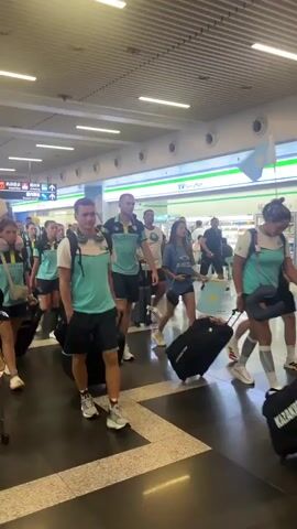 A group of people in matching teal and black athletic attire, some wearing travel pillows around their necks, are walking through an airport terminal. They are pulling luggage, mostly black suitcases on wheels. The people are diverse in gender and apparent age. One person in the foreground, a young woman with a travel pillow, smiles and makes a peace sign gesture towards the camera. Another person, a young man, also in the teal and black outfit, is walking ahead of her, pulling a black suitcase. The airport environment includes signs with Asian characters and a convenience store with a green awning. The floor is a polished dark tile. The lighting is bright, indicative of an indoor space with overhead lights.