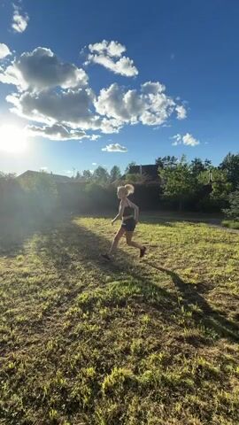 A woman with blonde hair tied in a ponytail is playing with a ball outdoors on a sunny day. She is wearing a dark sports bra and shorts. The grass is dry and patchy. In the background, there are trees and houses under construction. The sky is blue with scattered white clouds. The woman is in motion, kicking the ball and moving around the yard. There is another person's hand visible on the left side of the frame at the beginning of the video, possibly interacting with the ball or the woman.