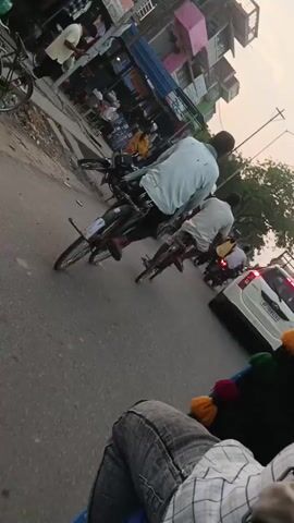 A video shows a street scene with several people and vehicles. In the foreground, the legs of a person wearing jeans and a plaid shirt are visible, seated in what appears to be a vehicle. Behind them, a rickshaw with colorful decorations is partially visible. The street itself is paved and appears to be busy.

Further into the scene, two men are riding bicycles. The man on the left is wearing a light-colored shirt and dark pants, while the man on the right is wearing a white shirt and dark pants. Both are cycling in the same direction.

A white SUV is stopped on the road behind the cyclists. The SUV has its brake lights illuminated and a license plate with "UP 09 D0923" visible. To the left of the SUV, a woman in a green saree is walking across the street. She is wearing jewelry on her wrists.

A motorcycle is parked on the left side of the road. Behind the motorcycle and the cyclists, there are several buildings with various colored facades, including pink and green. Some shops and stalls are visible along the street. Trees are present in the background. The lighting suggests it is daytime, possibly late afternoon, with a somewhat hazy sky.