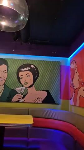The camera pans up and around to capture the interior of a vibrant and busy bar. In the foreground, a colorful booth with a yellow table and cushions in orange, red, and purple is visible. Above the booth, pop art-style paintings adorn the wall, depicting a couple with drinks and a woman’s profile. A disco ball hangs from the ceiling. As the camera moves, the bar area comes into view, with patrons seated at the counter, ordering drinks from the bartender. TVs are mounted on the ceiling displaying text. The bar itself is illuminated with colorful lights, and various pendant lamps hang above. Several tables are visible, with people seated. The overall atmosphere is lively.