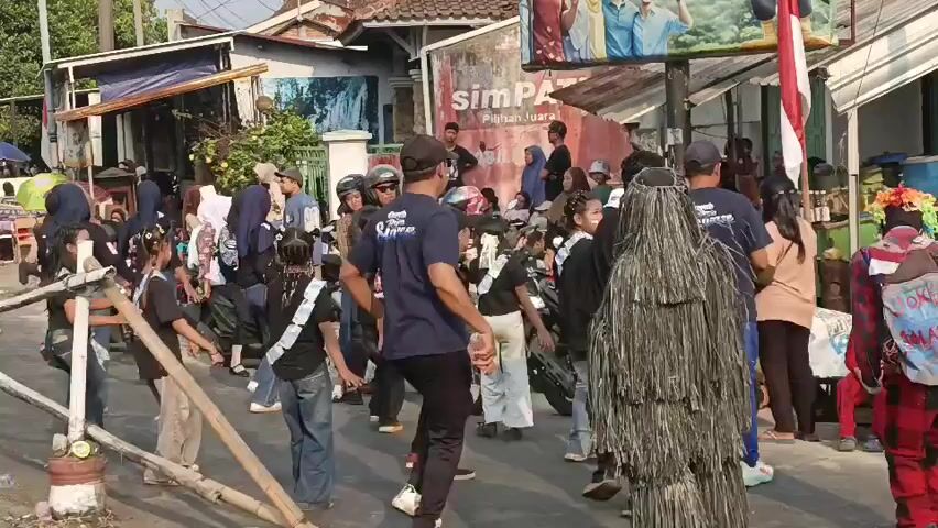 A street scene with a group of people, some wearing dark blue shirts with white writing, dancing or performing some synchronized movements. A person in a ghillie suit stands to the right, and another person in a jester-like costume is also visible. There are motorcycles and some onlookers in the background. A banner with Indonesian text is visible on a building. The ground appears to be paved.