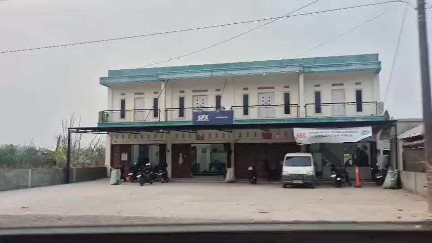 The video shows a street-level view of a two-story building with a light blue-green roof. The ground floor has several storefronts with open entrances, and motorcycles are parked in front of them. A white van is parked in front of one of the storefronts. Above the ground floor, the second floor has a balcony with railings. A sign with "SPX" in blue letters is visible above one of the storefronts. The building is situated next to a patch of dry grass and trees. The sky is overcast and the overall lighting suggests it might be daytime, possibly morning or late afternoon.