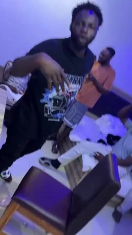 A video opens with a group of young men in what appears to be a dimly lit room with blue lighting. In the foreground, a man wearing a black t-shirt with a graphic design and grey shorts is captured in a low-angle shot. He is facing the camera, with his right hand near his waist and his left arm slightly extended. He has a beard and short, dark hair. Behind him, another man in an orange t-shirt and grey shorts is visible, holding a phone and looking at it. To the right and further back, a third man in a light blue t-shirt and white pants is reclining, with his head resting on his hand. A fourth man in a green t-shirt is also visible in the background. The room contains a bed with white bedding, and chairs with purple upholstery. The camera is at a slight tilt, and the lighting casts blue shadows. The footage appears to be filmed with a mobile device and shows some motion blur as the camera moves.