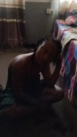 Here is a description of the video.

The video shows a woman sitting on the floor in a room. She is wearing a dark-colored cloth wrapped around her waist and her torso is bare. She has her right arm resting on her knee and her head is leaning on her hand. She appears to be looking at the camera. In the background, there is a bed with a colorful patterned bedspread and a man sitting on the bed. He is wearing a light-colored shirt and pants. The room appears to be dimly lit. A window is behind the man in the bed, and light from the window illuminates the space. The woman says, "Good morning everyone from me and my sibling."
