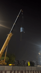 Construction crane lifts cylindrical object with blue surfaces at night