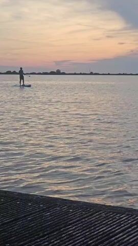 A person is paddleboarding on a body of water during sunset. The sky is filled with soft hues of orange and pink. The water is calm with gentle ripples. In the foreground, a dark, textured surface, possibly a dock or pier, is visible. The paddleboarder is a silhouette against the sky, moving away from the viewer. The horizon line is flat with distant landmass and some indistinct structures.