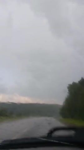 This is a video recorded from a vehicle driving on a wet road, likely in Kamennyi Kliuch, Russia. The scene is dominated by a cloudy, overcast sky, suggesting gloomy weather, possibly rain. The road is dark and appears to be wet, with visible ripples and splashes, indicating ongoing precipitation or recent heavy rain.

On either side of the road, there are dense green trees, forming a forest that lines the route. In the distance, a rolling landscape of trees creates a hazy, blurred backdrop, emphasizing the atmospheric conditions. The video captures the movement of the car, with the surrounding scenery appearing streaked and somewhat distorted, particularly due to raindrops on the windshield.

There are no people or prominent objects visible in the video, apart from the road itself and the natural landscape. The focus is on the experience of driving through this weather-affected environment. The overall mood conveyed is one of a somber, possibly isolated journey through a rain-swept rural or semi-rural area. The time of day is difficult to pinpoint precisely due to the heavy cloud cover, but it appears to be daytime. There is no visible text within the video.