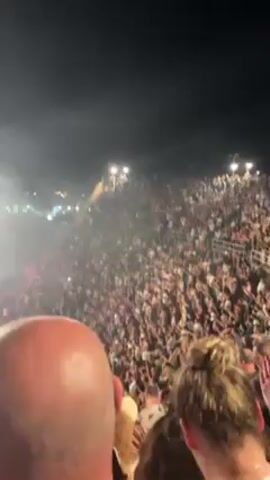 The video shows a large crowd of people at an outdoor concert at night. The crowd is packed and facing the stage, with many people holding up their phones to record. There are large screens on the stage displaying the performer. Some people are waving their hands in the air. The camera is held by someone in the crowd, and the view is partially obstructed by other people's heads and arms. The sky is dark.