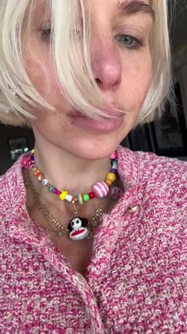 A fair-skinned woman with short, blonde hair is wearing a pink and white textured sweater and several necklaces. The necklaces consist of colorful beads, gold chains, and a pendant with a cartoonish head with red lips. The background is blurry, but includes framed pictures.