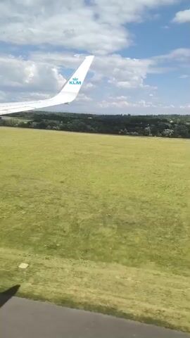The video shows a view from an airplane window during takeoff. The wing of a KLM airplane is visible in the upper left of the frame, along with the airline's logo. Below the wing, a large green field stretches towards a line of trees in the distance. The sky is blue with scattered white clouds. The plane accelerates down the grey asphalt runway, indicated by painted white lines.