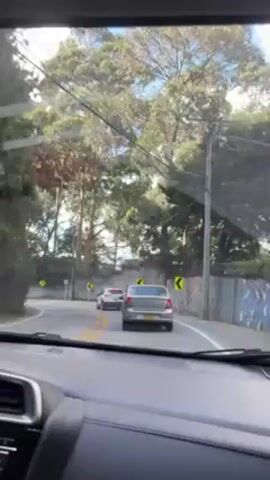 The video, shot from the driver's seat of a car, shows the vehicle traveling on a road that winds through a forested area that transitions into a more urban zone. Initially, the road is lined with trees and a stone wall, with a few other cars visible ahead. As the car progresses, buildings become visible, including some apartment complexes. A cyclist is briefly seen riding on the side of the road. The weather appears to be partly cloudy, with patches of blue sky visible above the trees and buildings. A sign indicating a speed limit of 30 is also visible.