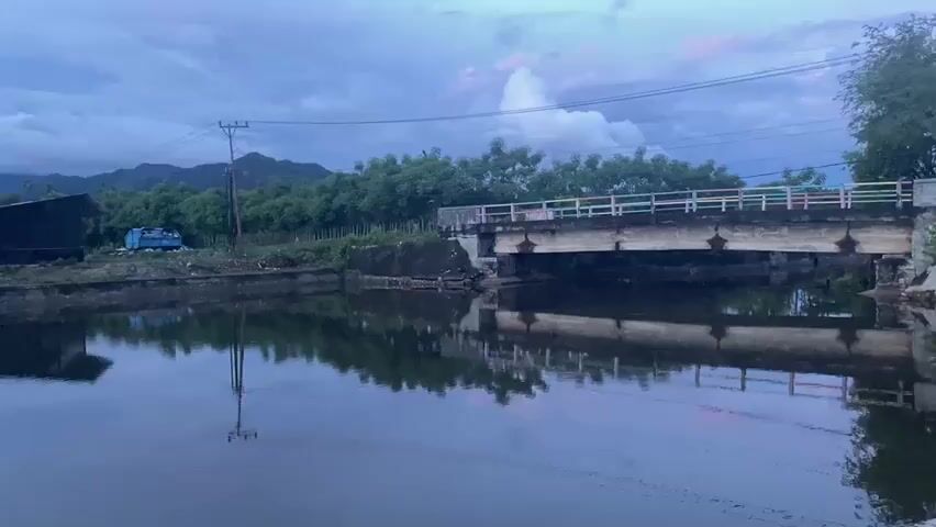 A tranquil body of water reflects a cloudy, twilight sky. In the background, a bridge spans the water, its railing a colorful pastel. Trees line the far bank, and beyond them, hazy mountains rise against the darkening sky. On the near bank, a small, dark building sits beside a light blue structure, possibly a water tank. A concrete barrier extends into the water from the left side of the frame, with blue plastic containers visible within it. The scene is quiet and still, with the water serving as a mirror to the muted colors of the evening.