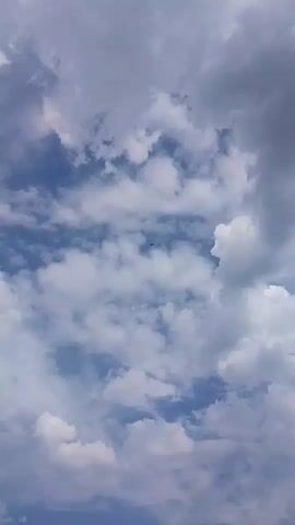 A video shows an airplane flying in the sky, surrounded by clouds. The airplane is visible as a small silhouette against the bright sky. The clouds are white and fluffy, with some darker clouds in the background. The sky is blue. The airplane appears to be moving from left to right across the frame.