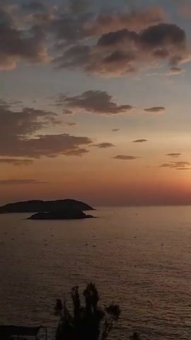The video shows a wide shot of a sunset over the ocean. The sun is a bright orange orb low on the horizon, casting a golden glow on the water and clouds. There are a few islands in the distance, their dark silhouettes contrasting with the vibrant sky. The water is calm, with small ripples reflecting the colors of the sunset. In the foreground, the tops of some trees are visible, silhouetted against the sky. The overall mood is peaceful and serene.