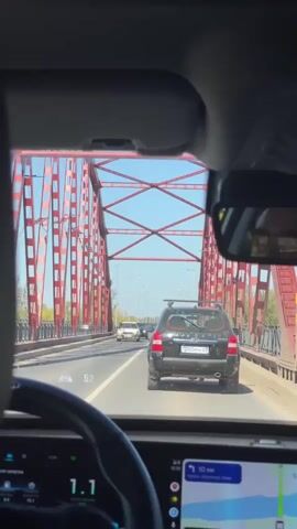 The video begins with a view from inside a car, looking forward through the windshield and side windows. The car is on a bridge with large red metal support structures visible on either side. There are other cars on the bridge, traveling in the same direction. The driver's side dashboard displays a speedometer reading of 1.1 and a navigation system with a map and route. As the car moves across the bridge, the view shifts, showing a section of the bridge's metal structure up close, then a wider shot of the landscape on the other side of the bridge, which includes a body of water and fields under a clear blue sky. The video is shot from the perspective of a passenger or driver, capturing the experience of crossing the bridge.