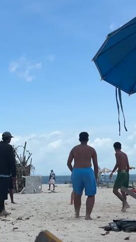 A group of men are playing soccer on a sandy beach under a cloudy blue sky. In the foreground, a man in blue shorts kicks a soccer ball towards the right of the frame. To his left, another man in a black t-shirt and shorts watches the ball. In the background, several other people are visible, including one in a black hat and jacket on the far left, and another in a white shirt and shorts in the distance. A makeshift structure made of branches and concrete blocks stands on the beach to the left of the main group of players. A blue umbrella is partially visible in the top right corner of the frame. The ocean is visible in the background.