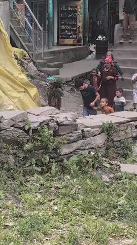 In this video, a group of people, including adults and children, are gathered near a stone wall. A young boy in a black shirt is holding the hand of another child in an orange shirt. Behind them, an adult woman in a burgundy headscarf and another woman in a green headscarf are present. A young child in a light-colored shirt is standing next to them, and another child in a patterned shirt is to the right. A dark-colored animal, possibly a dog or goat, is visible behind the children and in front of a yellow tarp. In the background, there are stairs leading up to buildings with shops. The environment appears to be outdoors, with overgrown grass and debris in the foreground.