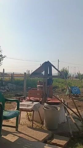 The video shows an outdoor scene with a well in the foreground. To the left of the well, there is a green chair and a table covered with a white cloth. A red chair is placed next to the well. A large white bucket is situated near the well. There are several wooden poles and tools leaning against the well structure. In the middle ground, there is a field with cultivated plants and a blue cart or wheelbarrow. A car is visible on a road in the background, along with power lines and trees. The sky is clear and blue, indicating a sunny day. The scene appears to be a rural or suburban backyard.