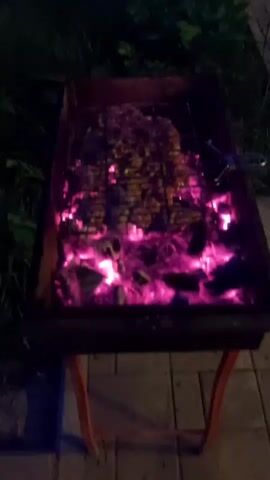 A close-up, overhead view shows food being grilled over hot coals at night. The food, likely meat, is arranged on a metal grill. The coals below glow a bright pinkish-purple, indicating intense heat. The grill is in a rectangular metal barbecue pit, which rests on an orange stand. The scene is set outdoors, with dark greenery visible in the background and a paved surface beneath the barbecue. The lighting suggests it is nighttime, with the grill's heat being the primary light source.