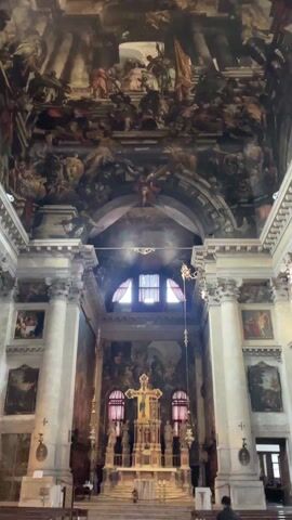 The interior of a church is displayed in the video. The primary focus is on the ornately painted ceiling depicting a multitude of figures, including what appears to be angels. The paintings extend down the walls and over an archway. Below, white pillars support the structure, and paintings are visible on the walls between the pillars. In the center is a golden altar with a cross. There is a window above the altar with red curtains. A voice of a child is heard in the background.