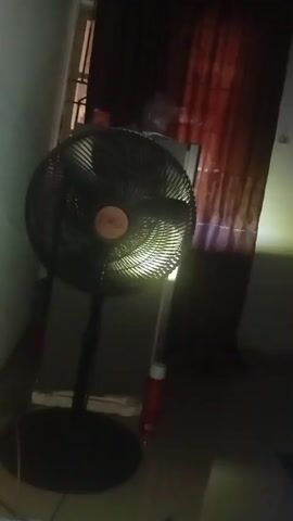 A black oscillating fan is in the center of the frame. It is standing on a black circular base. The fan has a black grate and a bronze-colored circular logo in the center. A light is shining on the fan from the right side of the frame, illuminating the grate. The fan is in a dimly lit room. To the left of the fan is a light-colored wall. Behind the fan, there is a gray object, possibly a cabinet or refrigerator, with a red can resting on it. Behind the gray object, there are dark red curtains. To the right of the fan, a beam of light is visible. The bottom of the frame shows a dark floor.