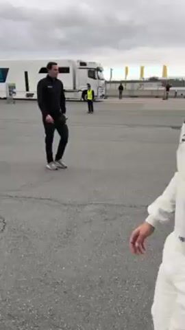 A man wearing a white and blue racing suit and glasses walks and turns to face the camera, giving a thumbs up. He smiles. He continues walking away from the camera. The scene is an outdoor parking lot or staging area with race team vehicles and tents in the background. It is a cloudy day.