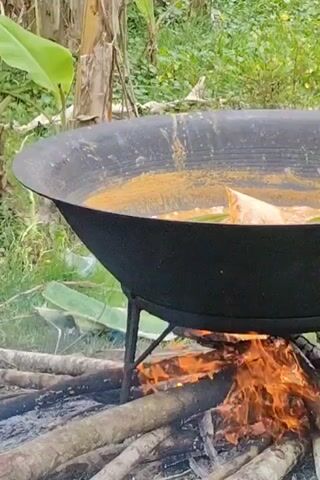 A large black wok-like pan sits atop a tripod stand over a wood fire in a grassy field. The pan contains liquid and food items, including what appears to be a banana leaf wrapping. The fire burns brightly underneath. The camera then pans to the right, showing scattered garbage including plastic bags, plastic bottles, banana leaves, and debris strewn across the grass. A chicken pecks at the ground amidst the trash.