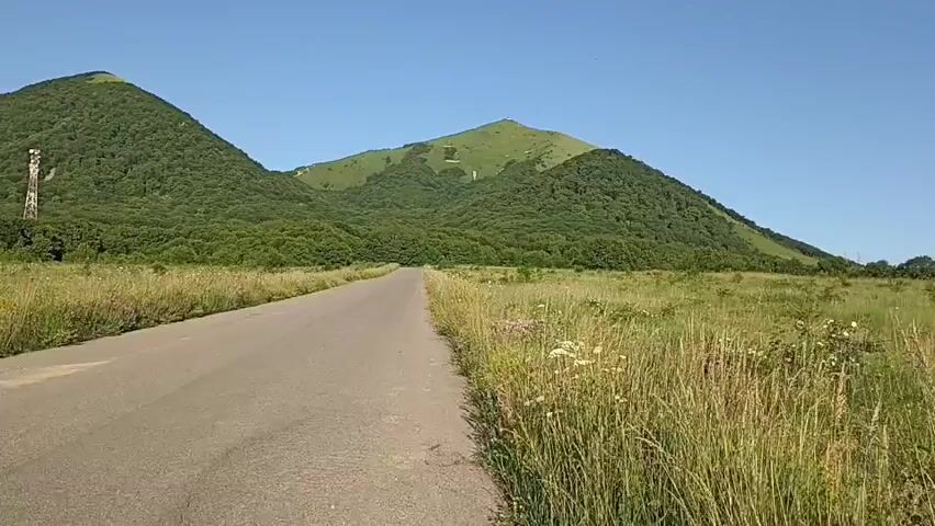 This video shows a view of a paved road stretching towards rolling green hills under a clear blue sky. The day appears sunny, with no clouds visible. The surrounding landscape is dominated by tall, dry grass on either side of the road, suggesting a warm season.

The hills themselves are heavily forested, with lush green trees covering their slopes. On the left hill, a prominent rocky outcrop is visible, and a tall, slender communication tower stands between the rocks and the trees. Further to the left, a white building complex can be seen nestled among the trees at the base of the hill. The right hill is also green and appears to have a lighter green patch, possibly a trail or a different type of vegetation, running down its side.

The overall impression is one of a peaceful, natural landscape. The road leads the viewer's eye into the distance, inviting exploration of the scenery. The presence of the communication tower and the building suggest that the area is inhabited or utilized, despite its natural beauty. The time of day seems to be midday or early afternoon, given the bright sunlight and the shadows cast. The location cues, combined with the name of the city, point to a scenic area in or near Lermontov, Russia. There is no discernible text within the video.