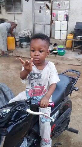A young child sits on a black motorcycle in front of a building. The child is wearing a white t-shirt with a cartoon car print and matching pants. The child faces the camera and makes hand gestures. Behind the child, an older woman wearing a headscarf can be seen lifting a yellow container. A man in a striped shirt walks in the background. The building has a door with containers stacked behind it and the number "4" is visible above the door. The surrounding area appears to be an outdoor courtyard.