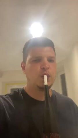 A man is shown from the chest up, wearing a dark t-shirt. He is holding a black hookah hose with a white mouthpiece to his lips and exhaling a large cloud of smoke directly towards the camera. The background is a lightly-colored wall with a light fixture on the ceiling. There is a piece of white material hanging on the wall behind him. The video appears to be a close-up, personal recording.