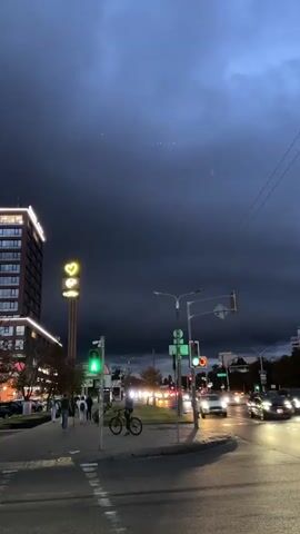 A time-lapse video shows a dark, cloudy sky over a city street at dusk. A tall building with illuminated windows stands on the left, and a yellow sign with a heart logo is visible. Traffic lights show green. Pedestrians walk on the sidewalk, some with bicycles. Cars drive on the street, their headlights illuminated. The sky darkens as the video progresses, with the clouds becoming more dense and covering the sky.